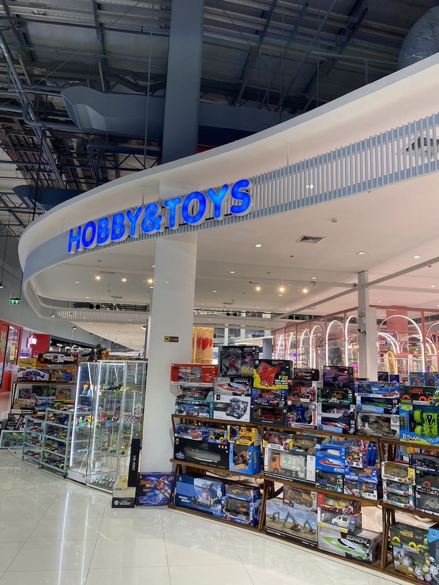 หน้าร้าน Hobby & Toys ป้ายชื่อร้านสีน้ำเงินบนผนังโค้ง มีตู้โชว์ของเล่นเรียงอยู่ด้านหน้า